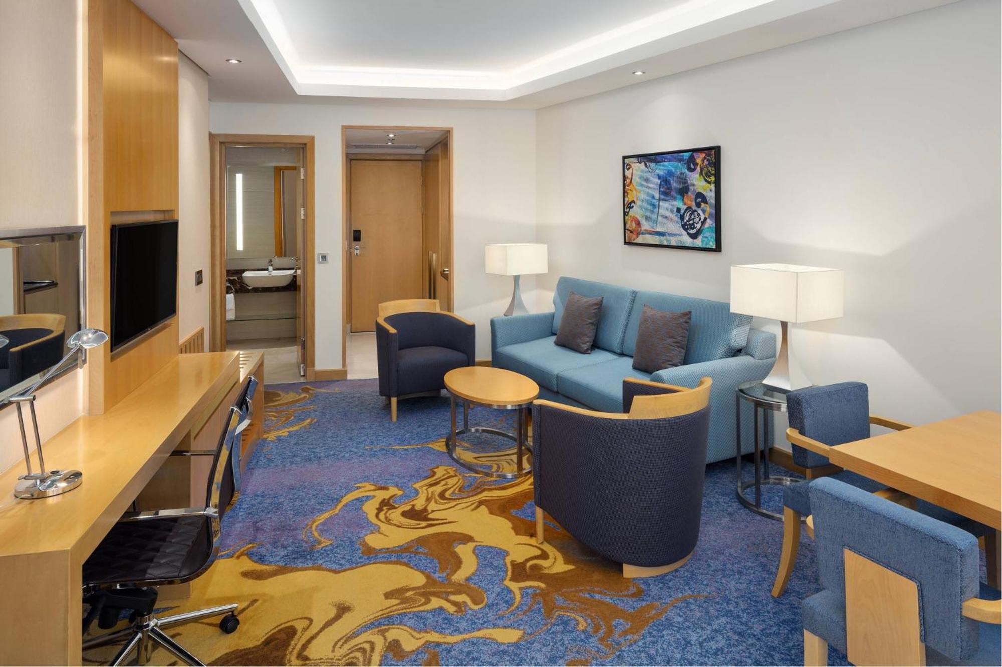 シェラトン メッカ ジャバル アル カアバ ホテル エクステリア 写真 The photo displays a modern hotel room or suite. The room features a comfortable seating area with a blue couch and two blue armchairs surrounding a small round table. A wooden desk with a black office chair is positioned against one wall, accompanie