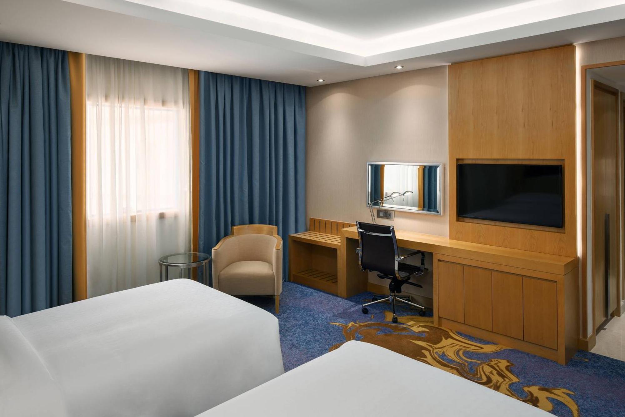 シェラトン メッカ ジャバル アル カアバ ホテル エクステリア 写真 The photo shows a hotel room featuring two beds with white linens. There are large windows covered with blue curtains that allow natural light to enter. On one side of the room, there is a wooden workstation with a chair and a mirror, along with a fl