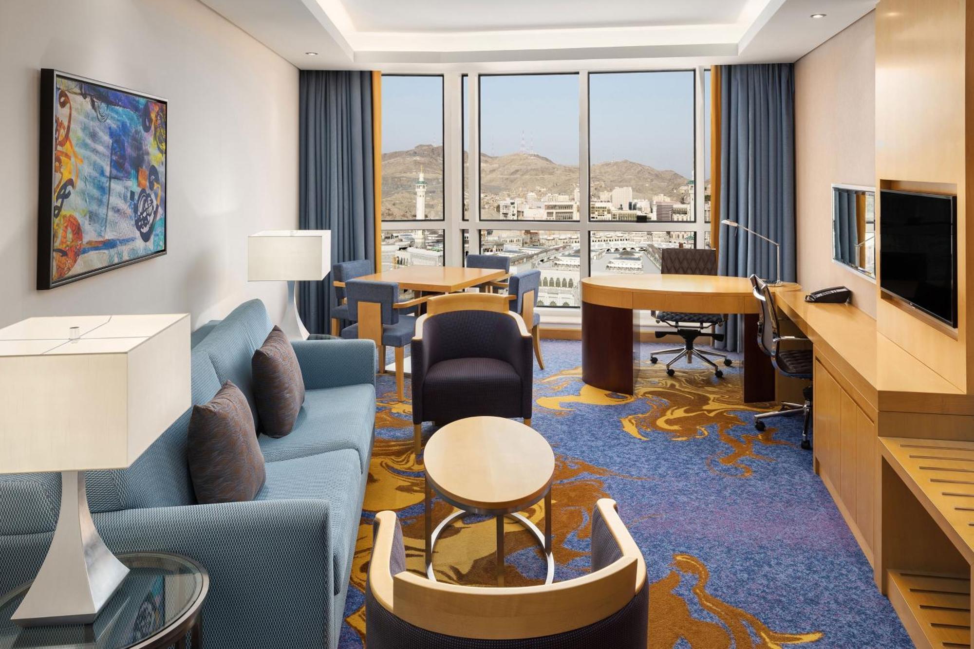 シェラトン メッカ ジャバル アル カアバ ホテル エクステリア 写真 The photo shows a well-decorated hotel room featuring a modern and comfortable interior. There is a cozy seating area with a blue sofa and armchairs, complemented by a round coffee table. A desk with a chair is also present, ideal for work. The room 