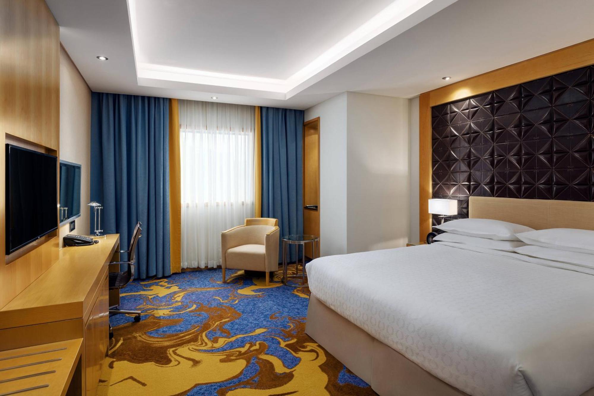 シェラトン メッカ ジャバル アル カアバ ホテル エクステリア 写真 The photo shows a modern hotel room featuring a comfortable double bed with a light-colored headboard. There's a large window dressed with blue curtains, allowing natural light to enter. The walls are painted in neutral tones, and one wall has a text